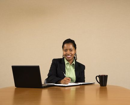 Businesswoman sitting at conference table with laptop computer and coffee cup smiling and writing in book.
