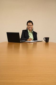 Businesswoman sitting at conference table with laptop computer and coffee cup smiling and writing in book.