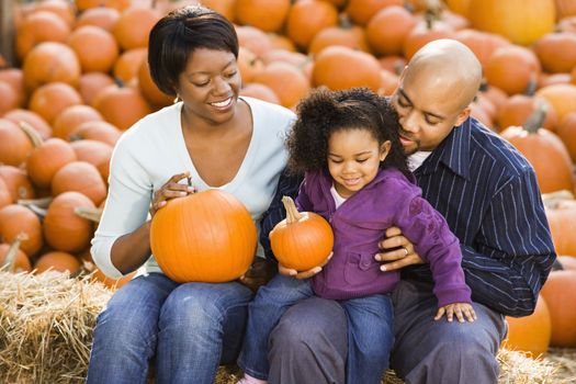 Happy smiling family sitting on hay bales and holding pumpkins at outdoor market.