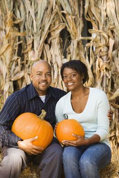 Happy smiling couple sitting on hay bales and holding pumpkins at outdoor market.