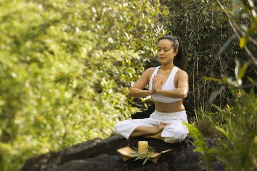 Asian American woman sitting on boulder in forest meditating with candle in Maui, Hawaii.