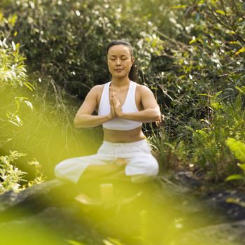 Asian American woman sitting on boulder in forest meditating with candle in Maui, Hawaii.