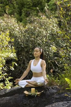 Asian American woman sitting on boulder in forest meditating with candle in Maui, Hawaii.