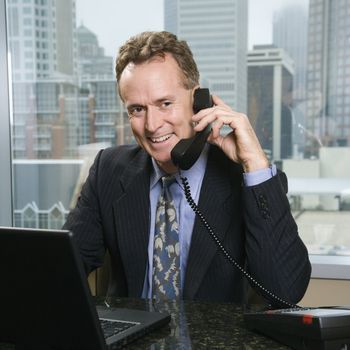 Middle-aged Caucasian male on phone in office with skyline in background.