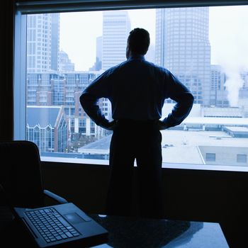 Middle-aged Caucasian male with hands on his hips in office with skyline in background.