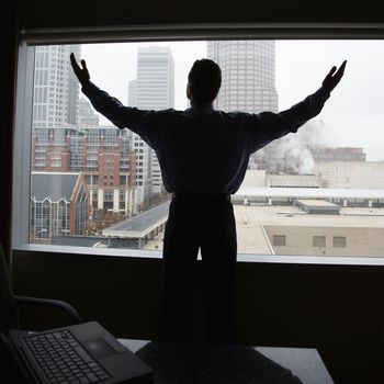 Middle-aged Caucasian male with raised arms in office with skyline in background.