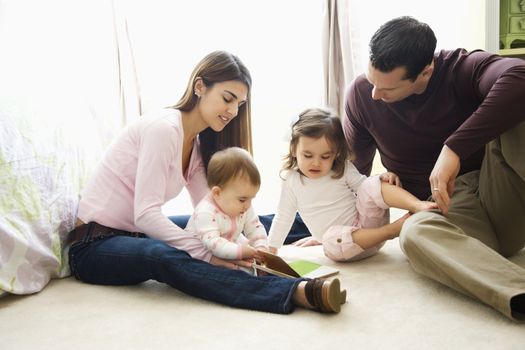 Caucasian girl children with mother and father sitting on bedroom floor looking at book.