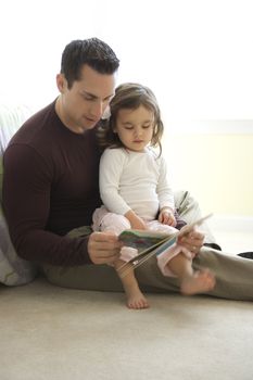 Caucasian father reading book to girl on lap sitting on floor.