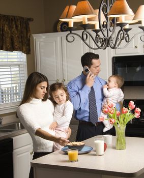 Caucasian mother and father in kitchen busy with children and cellphone.