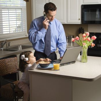 Caucasian father in suit talking on cellphone and using laptop computer with daughter eating breakfast in kitchen.