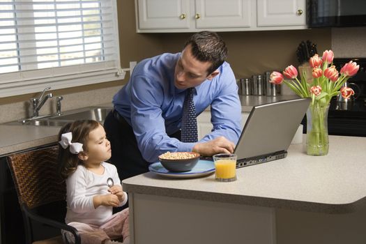 Caucasian father in suit using laptop computer with daughter eating breakfast in kitchen.