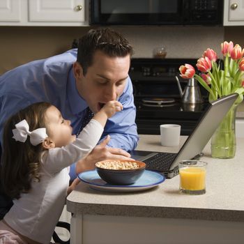 Caucasian father in suit using laptop computer with daughter feeding him breakfast in kitchen.
