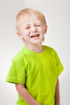 A boy of three years covering laughs (squinting) eye. Half-length portrait, arms down. Gray light background