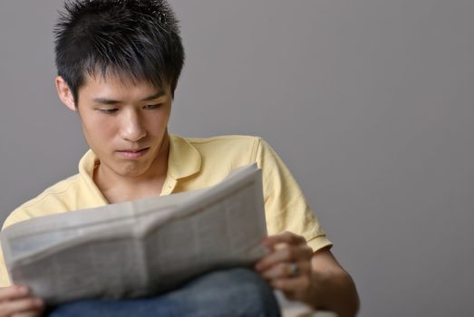 Asian young man reading newspaper over studio gray background.