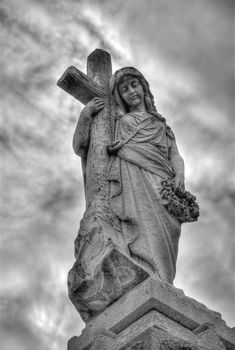 Madonna statue leaning against a cross adorning tomb in New Orleans, under dark moody skies.