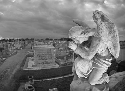 A praying statue kneels on top of a tomb in New Orleans under moody skies.  High point of view to contain the cemetery landscape in the background.