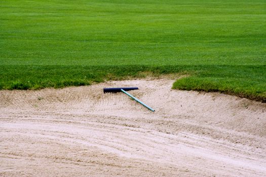 Sand trap on a golf course on the edge of the green with a rake laying in the sand