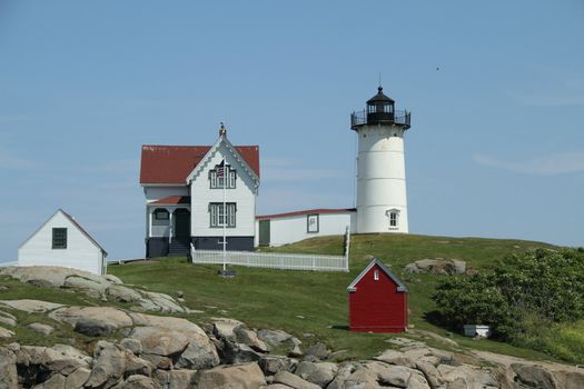 A lighthouse with out-buildings perched on a rocky landscape with green yard and blue, clear sky.