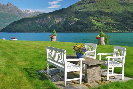 it is a garden place of a private cottage in the border of a fijord: the grass with a table and chairs decorated with flowers, is a part of norwegian lifesile; beside fjord there are mountains covered with green woods, and with snow in top 