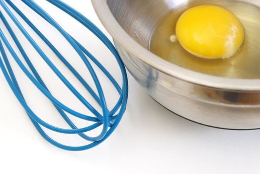 A raw egg in a mixing bowl with a blue whisk beside it.