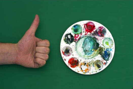 Human hand with thumb up positioned next to the artists palette with multiple paint colors