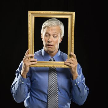 Caucasian middle aged businessman holding empty picture frame in front of face making expression.