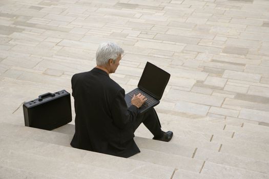 Back view of Caucasian middle aged businessman typing on laptop with briefcase sitting on steps outdoors.