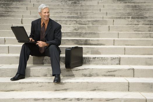 Caucasian middle aged businessman sitting on steps outdoors with laptop and briefcase.