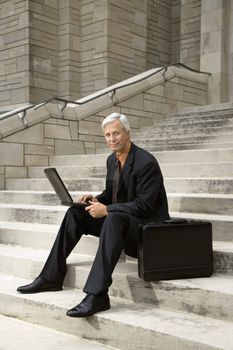 Caucasian middle aged businessman sitting on steps outdoors with laptop and briefcase.