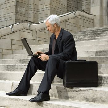 Caucasian middle aged businessman sitting on steps outdoors with laptop and briefcase.