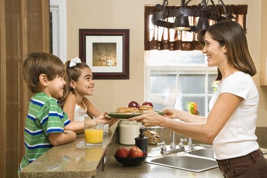 Hispanic mother handing healthy breakfast to young children in home kitchen.