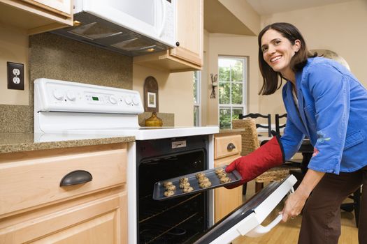 Hispanic mid adult woman putting cookies into oven and smiling at viewer.