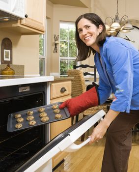 Hispanic mid adult woman putting cookies into oven and smiling at viewer.