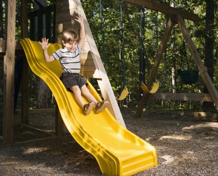 Hispanic boy sliding down outdoor slide with arms raised above head.