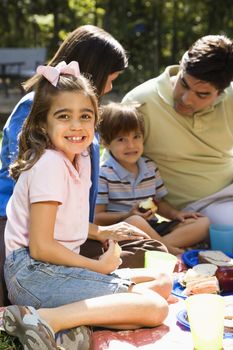 Hispanic girl smiling at viewer with family picnicking in the park.