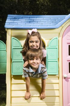 Hispanic boy and girl posing in window of playhouse and smiling at viewer.