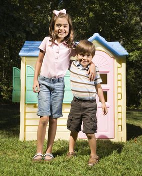 Hispanic boy and girl in front of outdoor playhouse smiling at viewer.