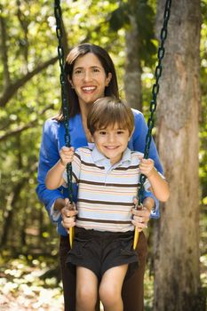 Hispanic mother pushing son on swing and smiling at viewer.