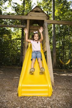 Hispanic girl sliding down outdoor slide with arms raised above head.