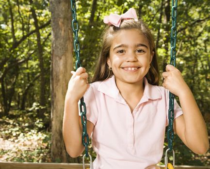 Hispanic girl sitting on playground swing smiling at viewer.