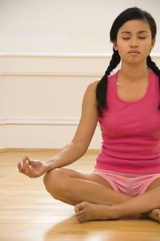 Young woman sitting on floor meditating in yoga lotus pose with legs crossed and eyes closed.