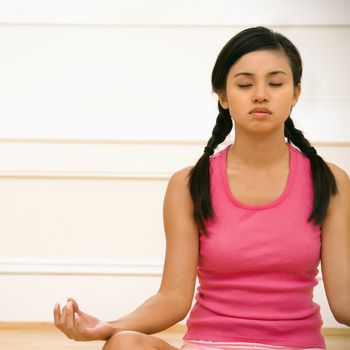 Young woman sitting on floor meditating in yoga lotus pose with legs crossed and eyes closed.