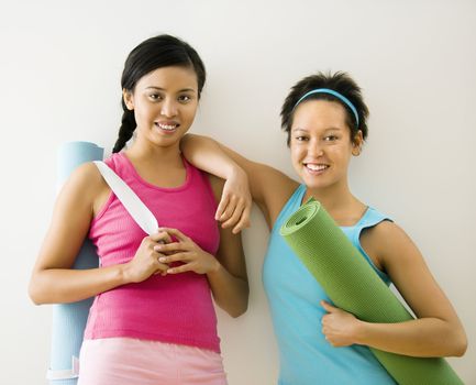 Two young women standing in workout clothes holding yoga mats and smiling.