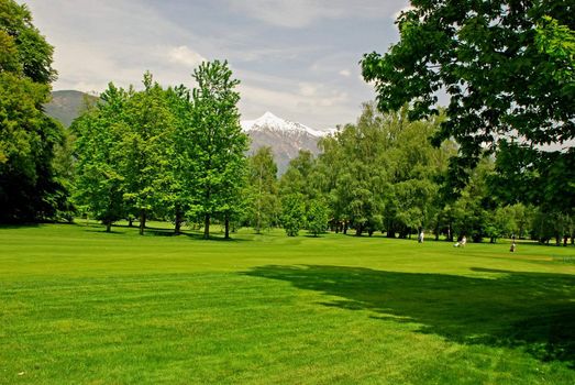 There is very beautiful golf park in Ascona situated in the border of the Lake Maggiore. Some people are preparing to play golf surrounded by park trees, there are alp mountains and the sky at the backgroung