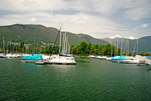 There is a sailing port and yacht club in Ascona, in Lake Maggiore. Behind yachts, there is a park,  at the background ther is a part of Locarno city and Alps with snow covered peaks of   Trosa and Madone mountains and cloudy sky