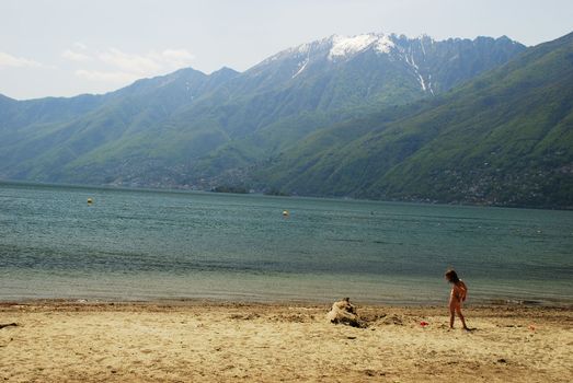 There is a Ascona beach in Lake Maggiore in the middle of May, there is a child playing in the beach, there is a beutiful view in the lake, porto Ronco and  Brissago situated in the border of the Lake, beutiful Leone hill still covered with snow and the cloudy sky