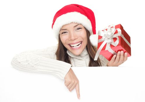 Christmas woman leaning over billboard sign. Pointing down holding gift showing big toothy smile. Caucasian / Asian woman wearing Santa hat and winter sweater isolated on white background.