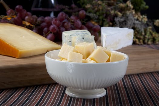 Cheese cubes in a bowl on a table with cheese  and grapes in the background.