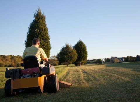 Middle aged man on zero turn mower cutting grass on a sunny day with the sun low in the sky