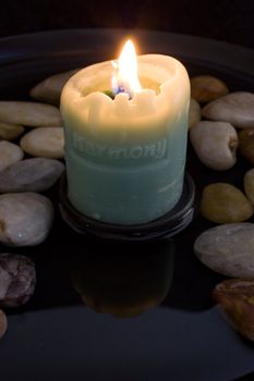 Lit "harmony" candle reflected in a peaceful pool of water surrounded by smooth stones.